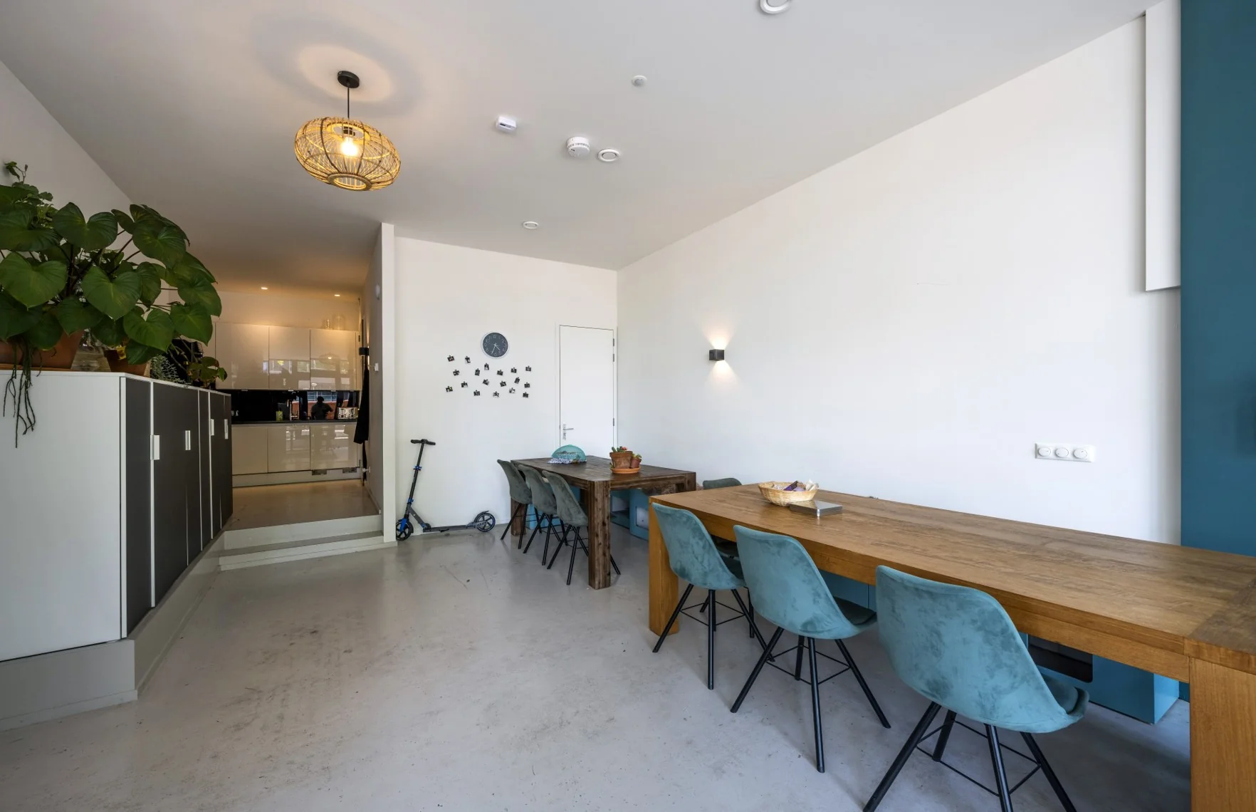 Moderne eetkamer met twee houten eettafels, blauwe stoelen en open doorgang naar een witte keuken.